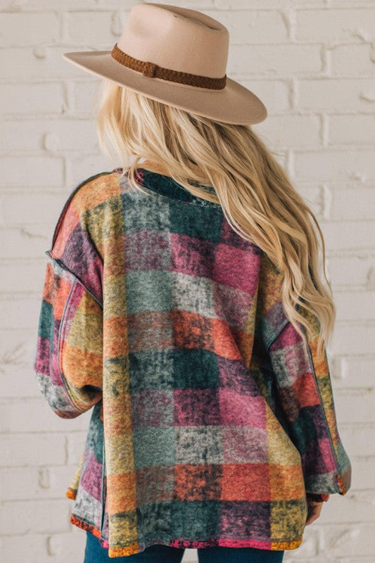 A person wearing a Mustard Red plaid button Shacket Cardigan Sweater made of 100% polyester over a white henley top and dark jeans, standing against a white brick wall.