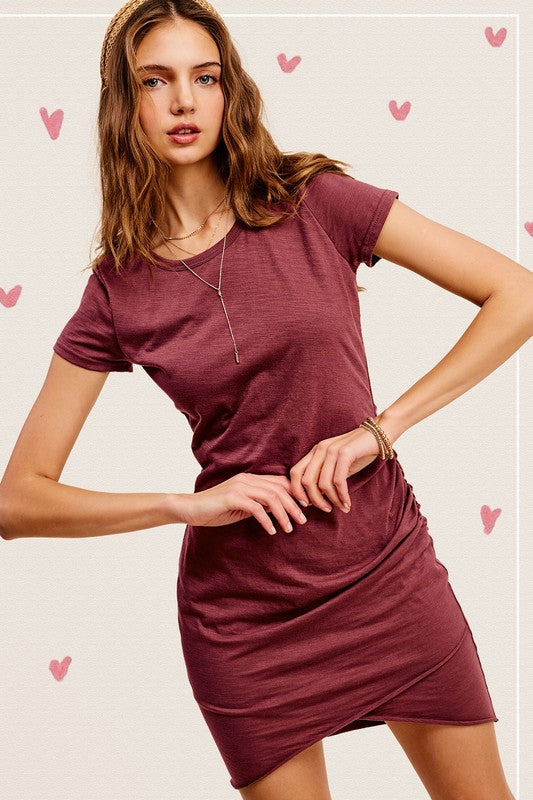 A woman with long hair is wearing a Soft Ruched Round Neck Short Sleeve Mini Dress in maroon, standing with one hand on her hip against a neutral background adorned with ribbon details.