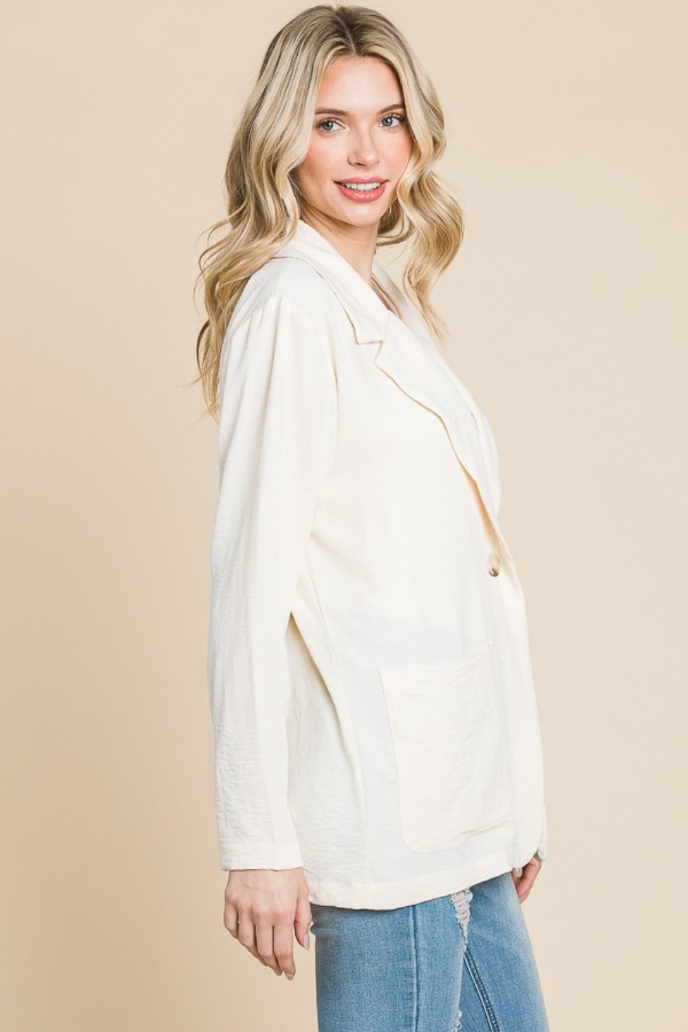 A person with long hair wearing the Culture Code One Button Long Sleeve Blazer with Pockets in light cream stands against a beige background, smiling. The blazer's tailored fit perfectly complements the white top and blue jeans, creating a versatile look for any occasion.