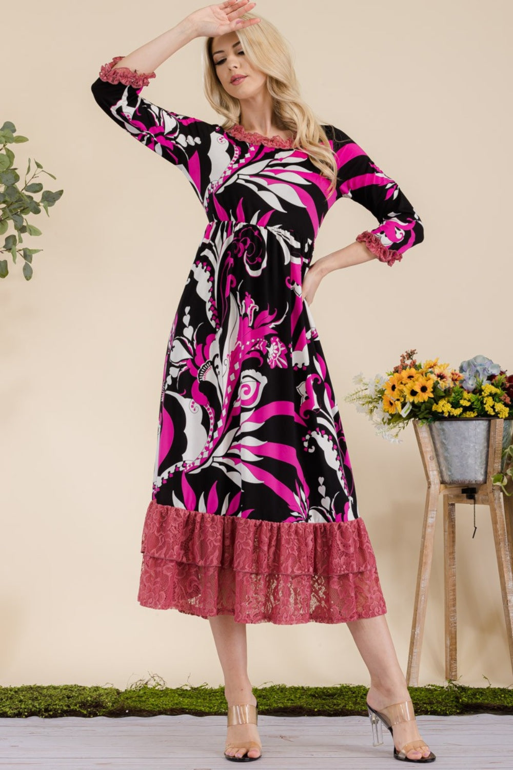 A woman wearing the Celeste Full Size Paisley Print Lace Ruffled Midi Dress poses gracefully for the camera indoors, standing beside metal planters filled with flowers.