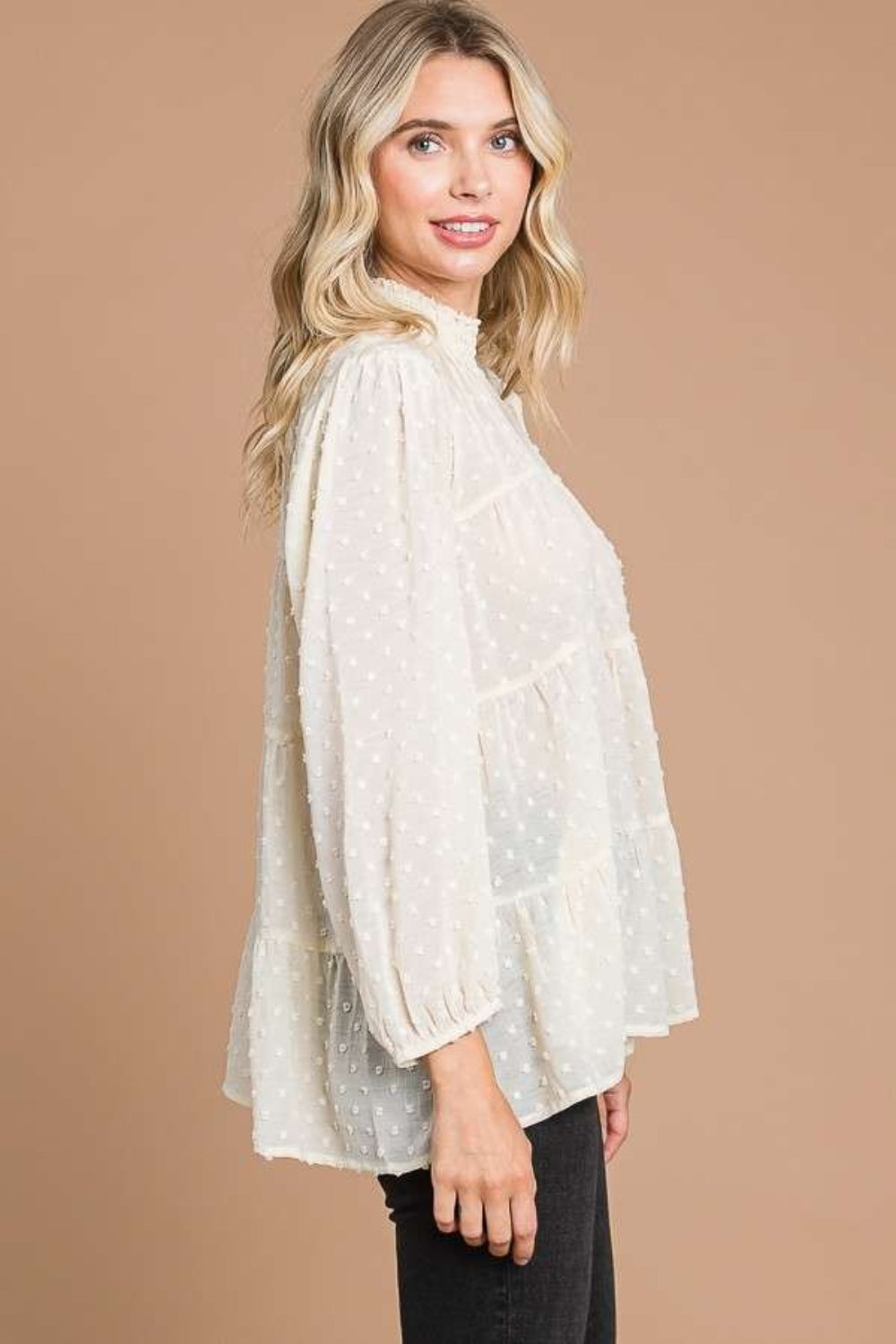 A woman with blonde hair stands against a plain brown background, exuding feminine fashion elegance in a loose, long-sleeved Culture Code Full Size Swiss Dot Smocked Mock Neck Blouse.