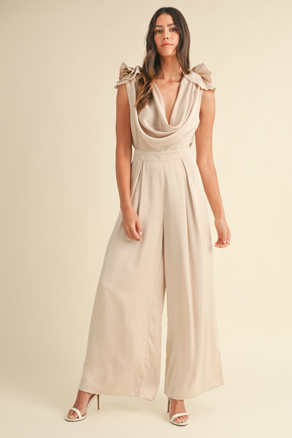 A woman stands wearing the MABLE 3D Floral Applique Deep Cowl Neck Jumpsuit, which showcases a light beige color with a draped neckline and ruffled sleeves. Featuring a wide leg silhouette, the outfit flows elegantly. She has long hair and is wearing high heels.