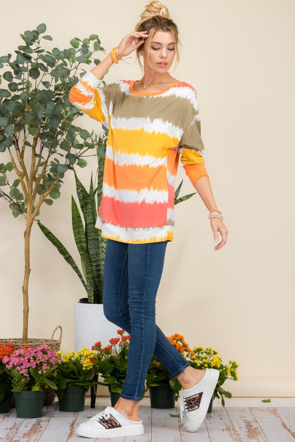 Wearing the Celeste Full Size Striped Long Sleeve T-Shirt paired with jeans, a person smiles while standing against a light background adorned with plants, exuding a casual yet stylish look.