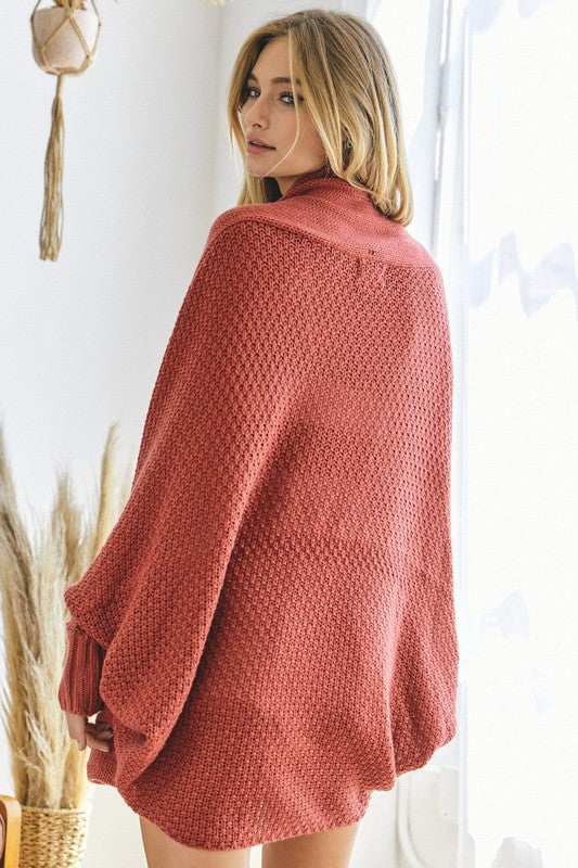 A person stands against a plain background, wearing a Pattern Knit Dolman Sleeve Solid Slouch Cardigan in textured blue over a white sweater with distressed white shorts, hands in cardigan pockets.