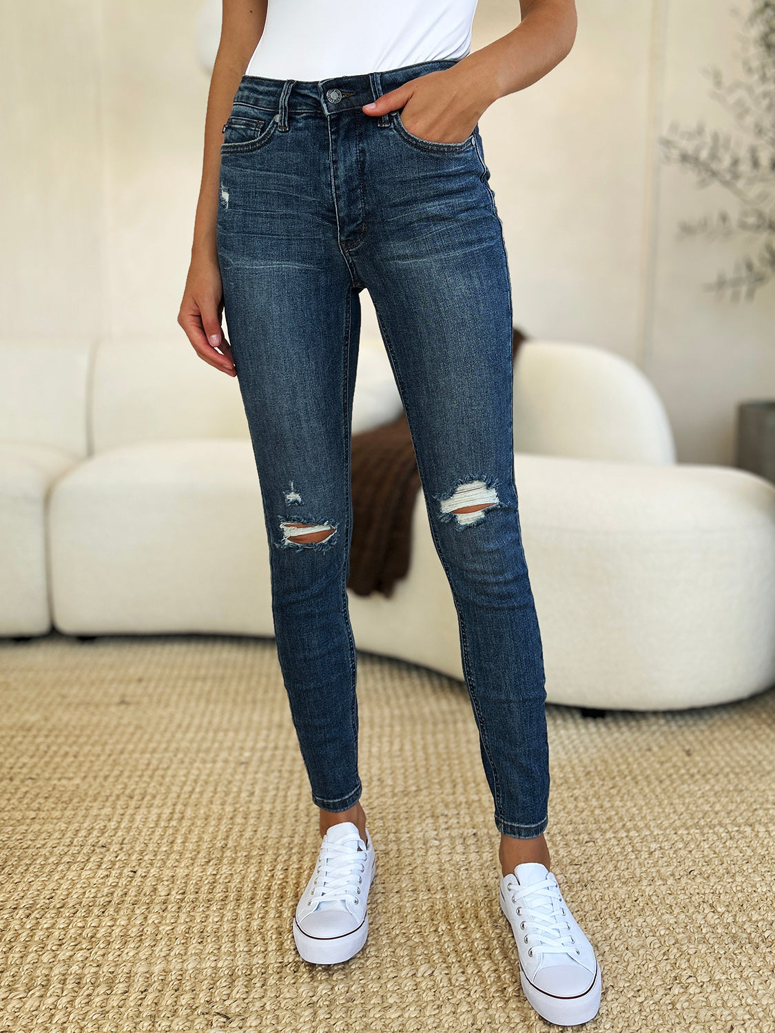 Person standing indoors wearing Judy Blue Full Size Mid Waist Distressed Slim Jeans, a black top, and black sneakers with white laces. There is a white sofa and a textured rug in the background.