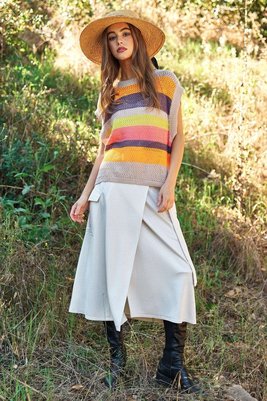 A woman outdoors wearing a Crochet Multi Striped Pullover Knit Sweater Vest and white pants, with her arms raised and eyes closed.