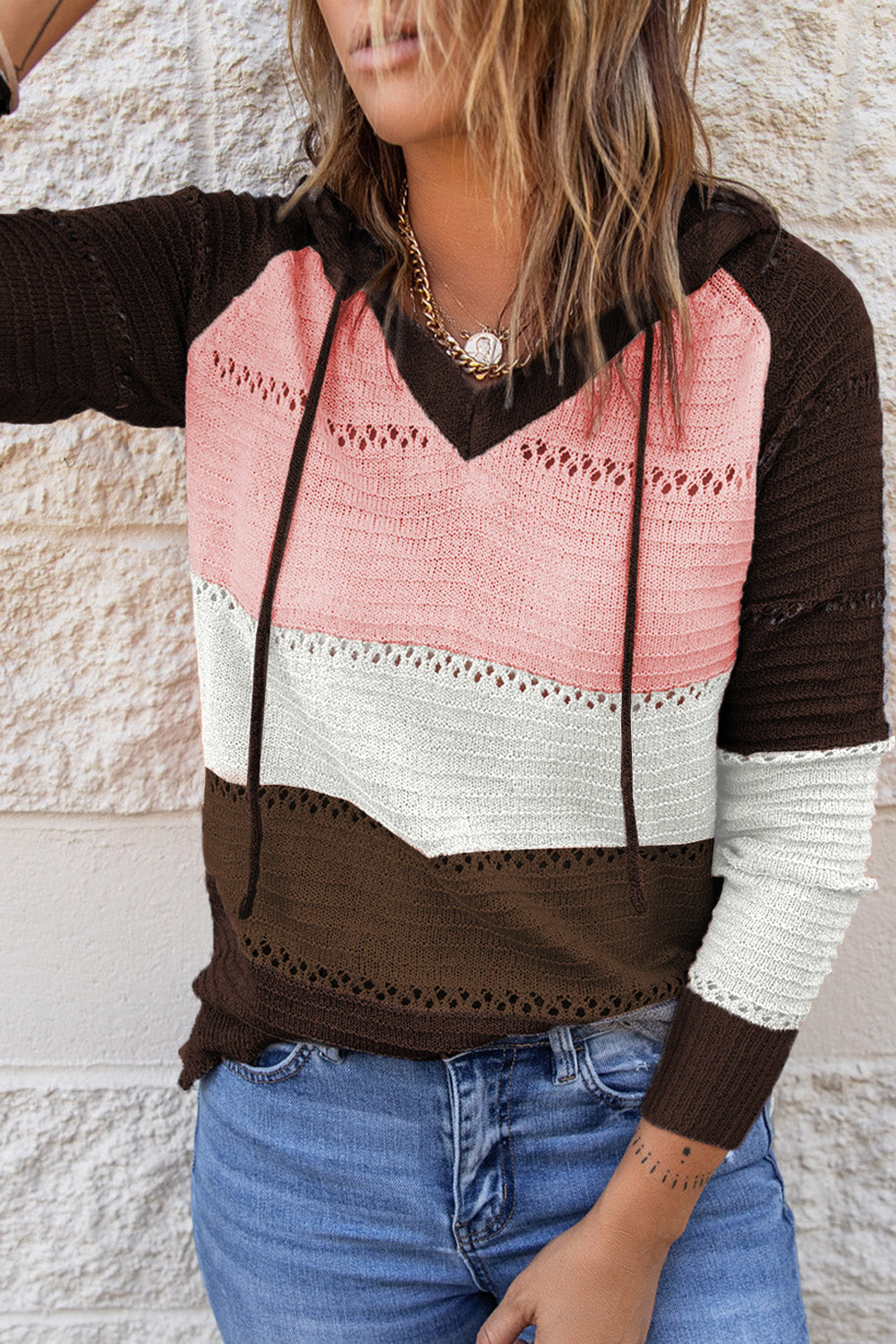 A person stands with their back to the camera, adjusting the hood of a Brown Beach Bonfire Knitted Hoodie featuring dark brown, pink, white, and light brown stripes. Perfect for a casual style, they are wearing distressed blue jeans and showcasing the laid-back vibe of lightweight hoodies.