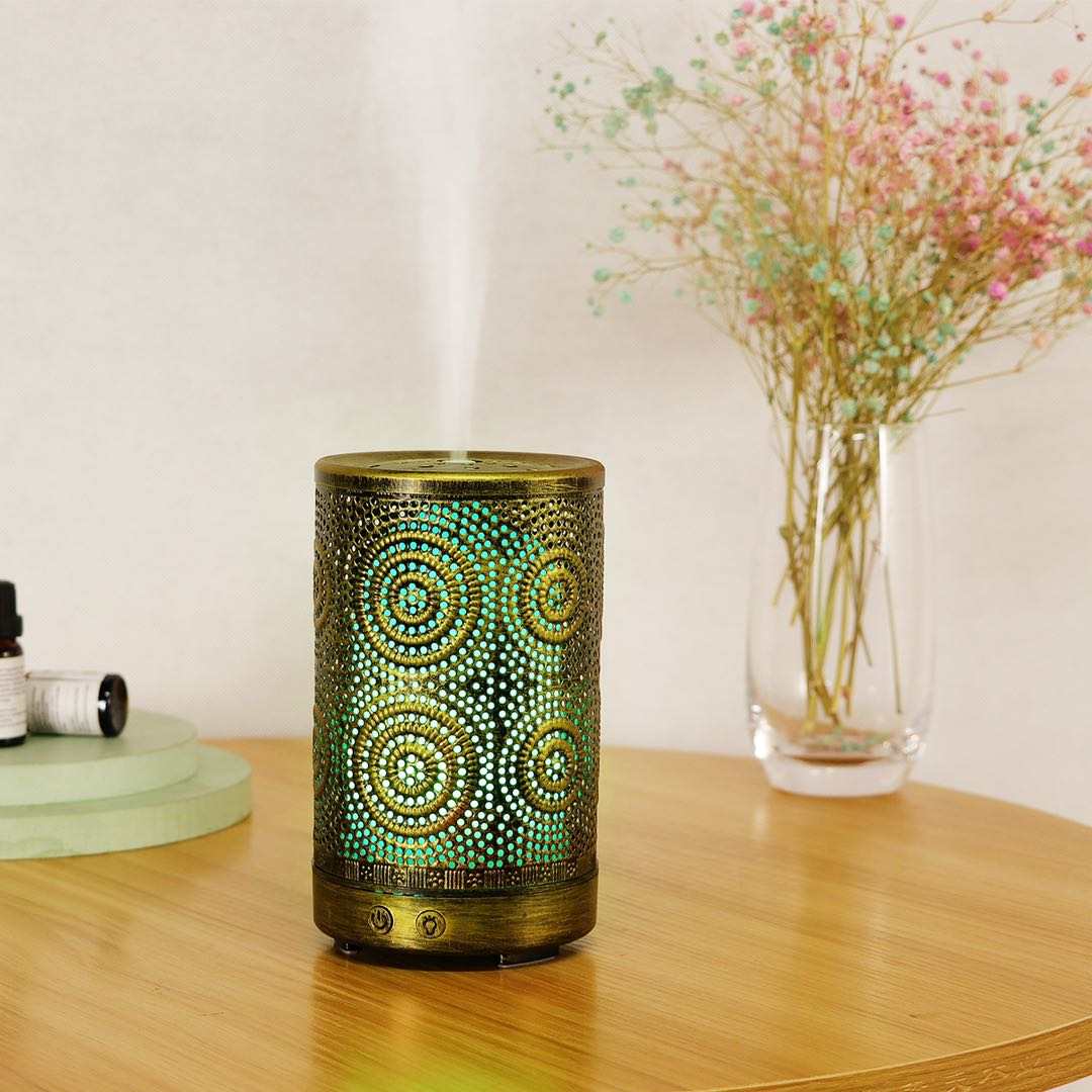 A chic Color Changing Aromatherapy Oil Diffuser with a floral motif rests on a wooden table, softly releasing mist enhanced with essential oils. In the background, a vase displays vibrant flowers, crafting an ideal aromatherapy atmosphere.