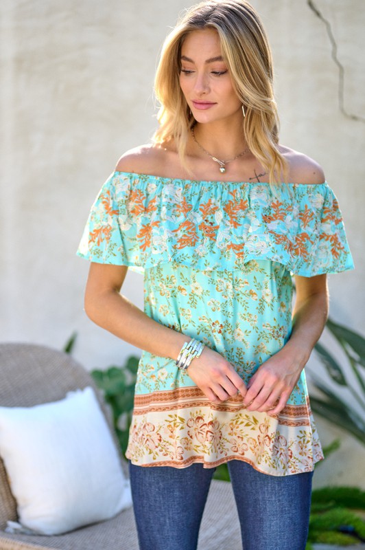 A woman with blonde hair is smiling while standing outdoors, wearing the Printed Off Shoulder Smocked Top paired with jeans.