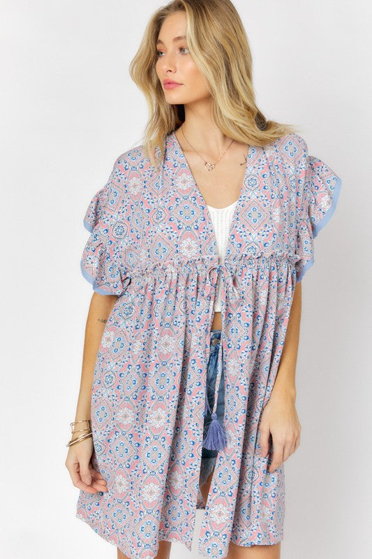 A woman stands against a white background wearing the Printed Short Sleeve Ruffle Kimono over a white top and denim shorts, effortlessly blending style and comfort.