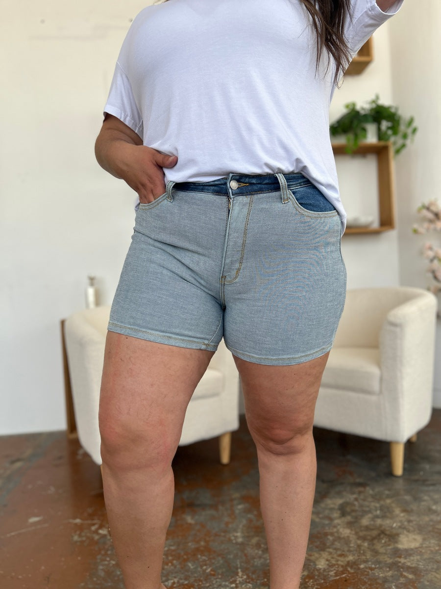 A person models indoors beside a beige chair and a plant, sporting a white t-shirt with the Judy Blue Full Size Color Block Denim Shorts for an effortlessly bold, eye-catching look.