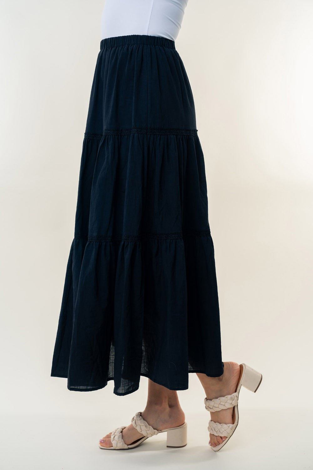 A long, White Birch High Waisted Tiered Maxi Skirt in navy blue, offering a versatile piece perfect for a bohemian look. It's worn by a person paired with white heeled sandals. The background is plain and light-colored.