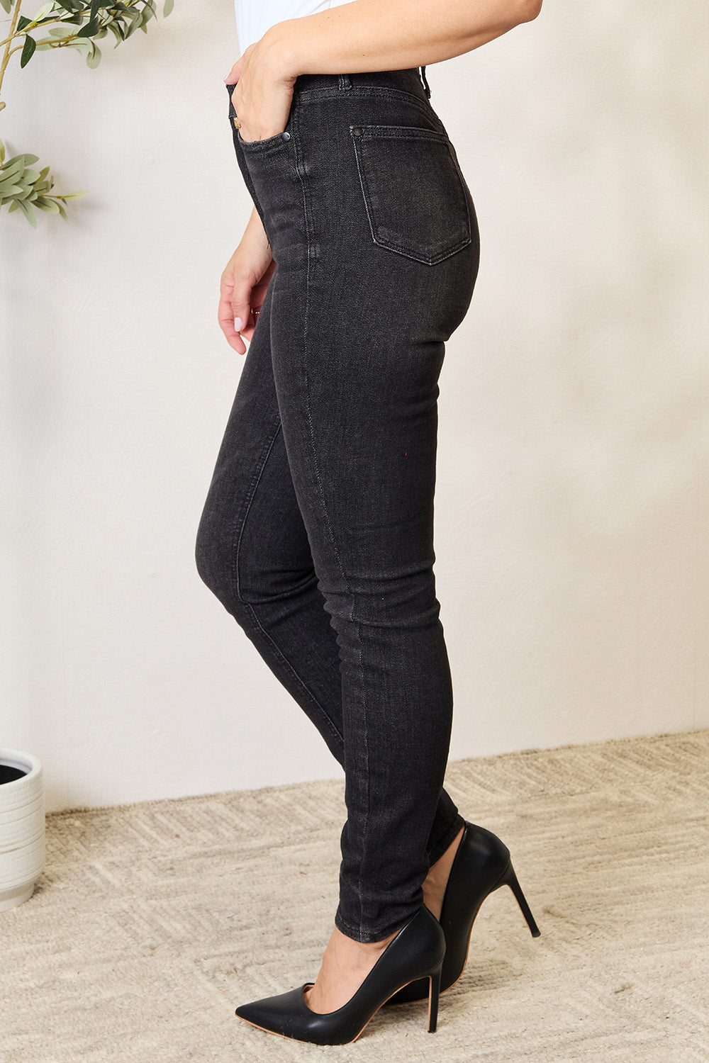 A person wearing a white top and Judy Blue Full Size Tummy Control High Waist Denim Jeans, made with pocketed stretch fabric, stands with one hand on their leg near a potted plant.