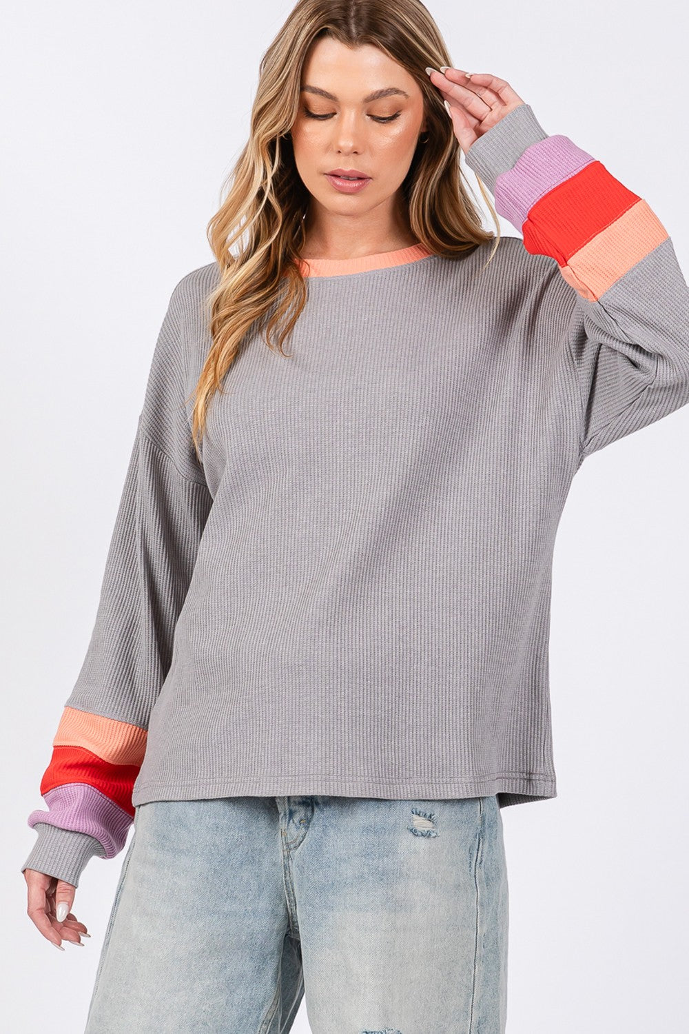 A woman wearing the SAGE + FIG Color Block Waffle Knit T-Shirt stands with one hand on her gray hat. The casual top, adorned with colorful block raglan sleeves, pairs effortlessly with denim shorts for a chic look.