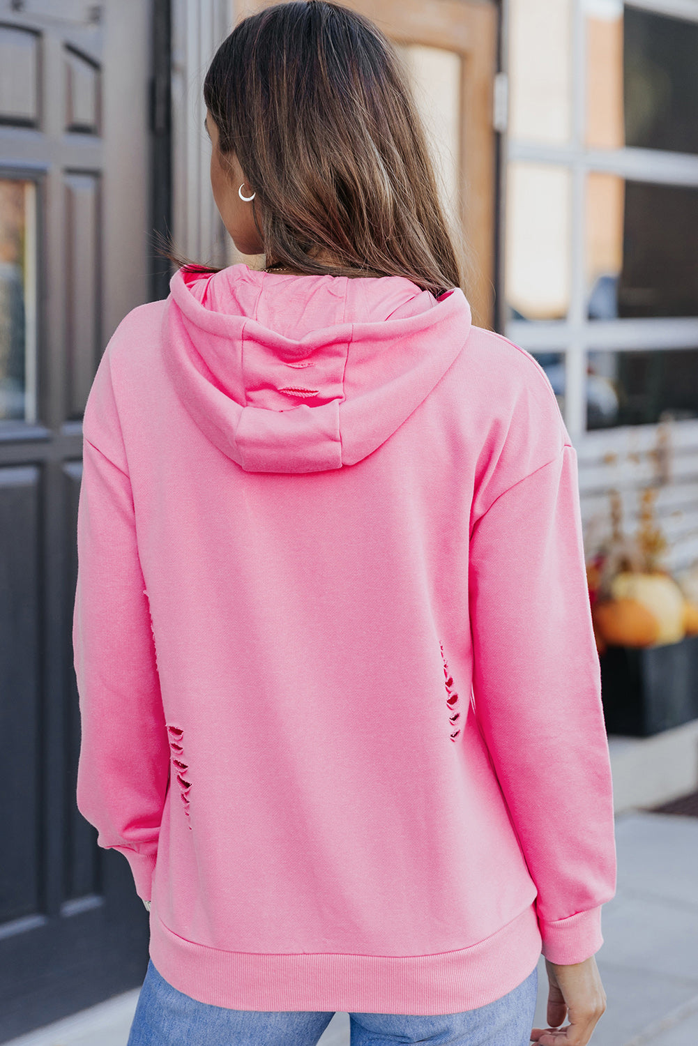 A person wearing a Pink Solid Ripped Hooded Sweatshirt with a Kangaroo Pocket is standing in front of a door.