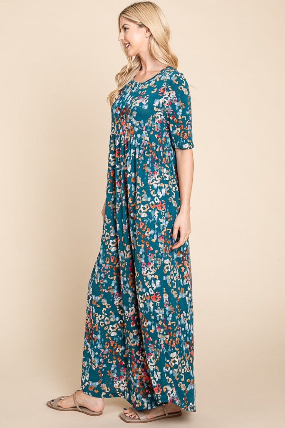 A person wearing a BOMBOM Printed Shirred Maxi Dress, featuring an eye-catching teal floral pattern and short sleeves, stands against a beige background.