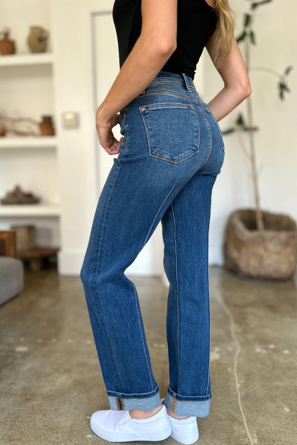 Two women stand side by side indoors, both wearing black tank tops, Judy Blue Full Size High Waist Front Seam Detail Straight Jeans, and white shoes. They are smiling and posing for the camera. A plant and shelves with decorations are in the background.