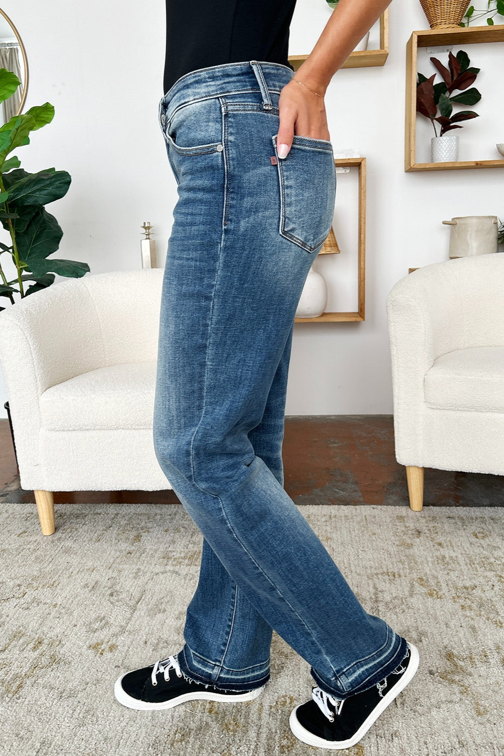 A person standing indoors, wearing a pair of Judy Blue Full Size Mid Rise Release Hem Jeans and black sneakers, holding the loose fabric of the jeans on one side.