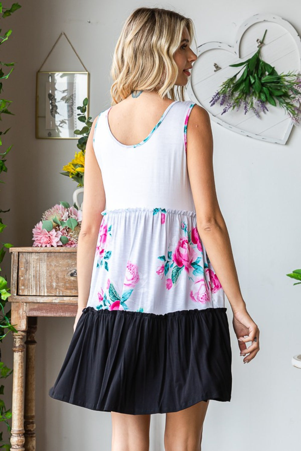 The woman wears the Heimish Full Size Scoop Neck Frill Tiered Dress, showcasing its versatile design with floral and black color blocks. She is indoors, beside a wooden table adorned with plants and wall art.