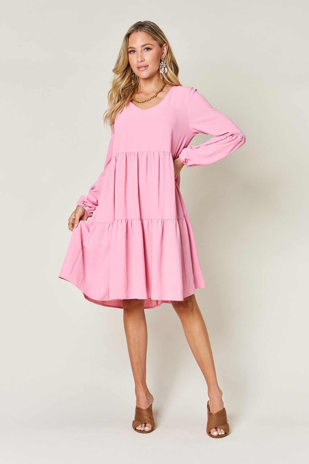 A woman stands wearing the Double Take Full Size V-Neck Balloon Sleeve Tiered Dress with Pockets in pink. Crafted from high stretch yarn, this dress combines comfort and style effortlessly. She has long, wavy hair and accessorizes with hoop earrings and a necklace. This imported garment is easy to care for – simply machine wash cold to keep it looking fresh.