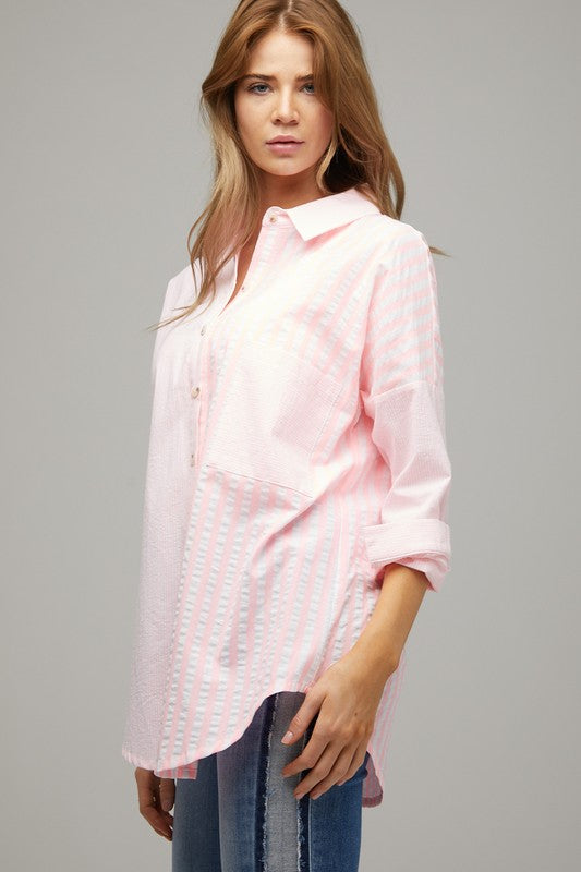 A person wearing a light pink Stripe Button Down Long Sleeve Shirt with rolled-up sleeves and jeans stands with their hands behind their head against a plain background.