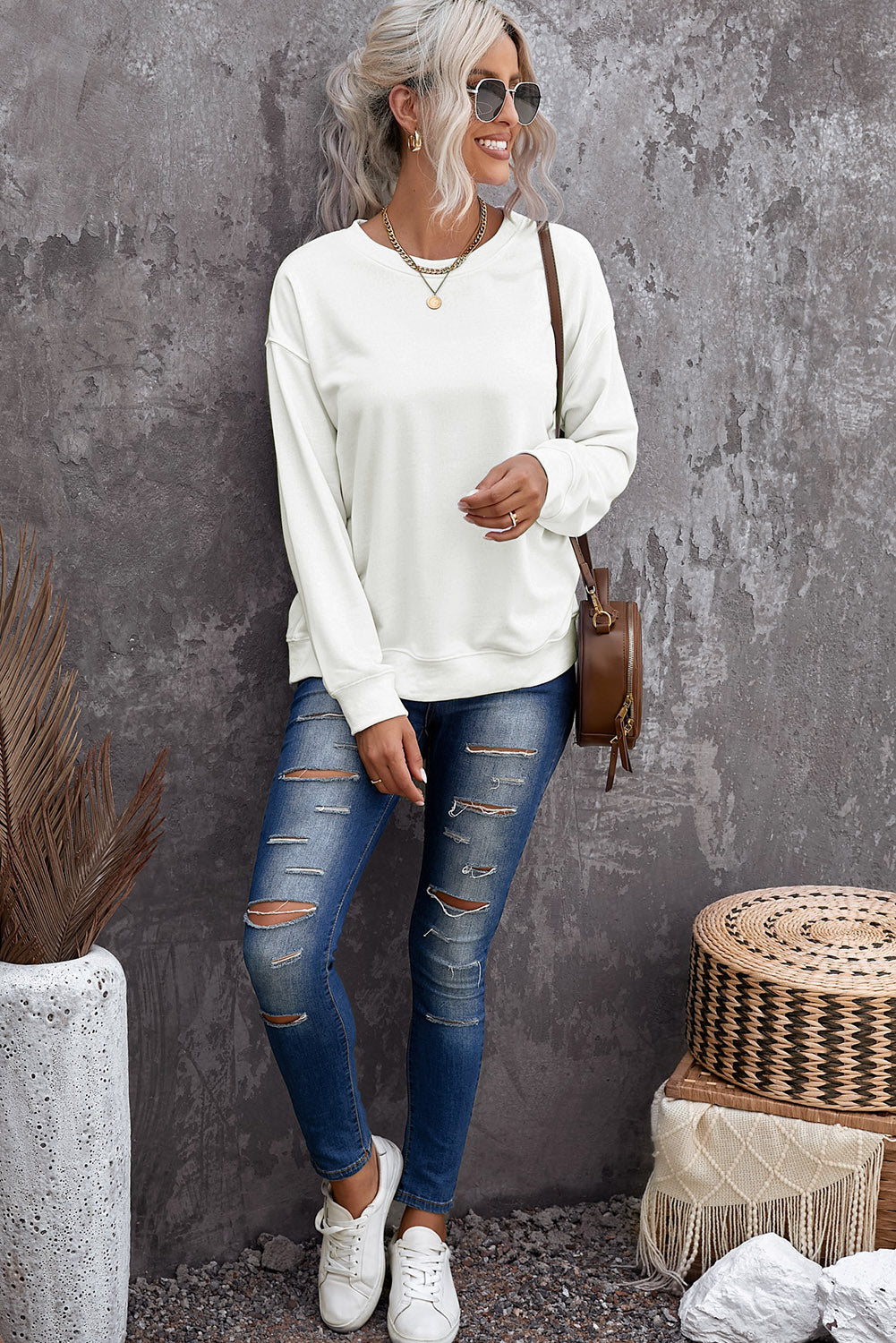 A woman stands in a festive room decorated with a Christmas tree and stockings, showcasing her winter fashion in a beige plain crew neck pullover sweatshirt and jeans.