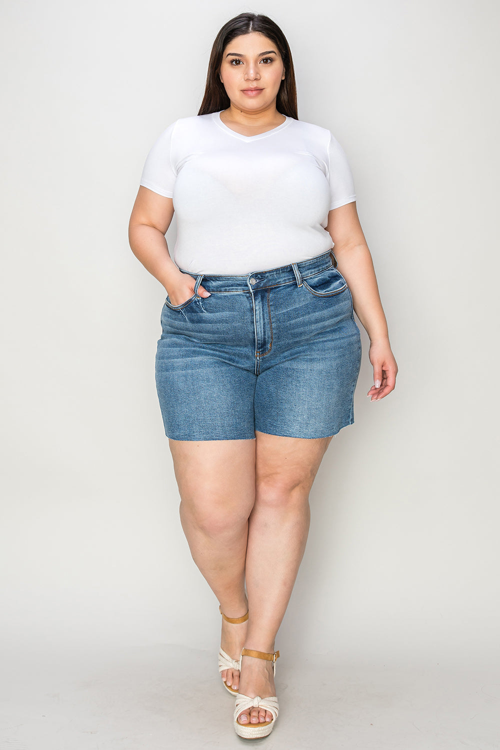 A woman in a white short-sleeve top and Judy Blue Full Size High Waist Slim Denim Shorts stands against a plain backdrop. She is smiling with her hands resting on her hips, wearing white open-toe wedge sandals that emphasize the quality denim fabric of her high-waisted design.