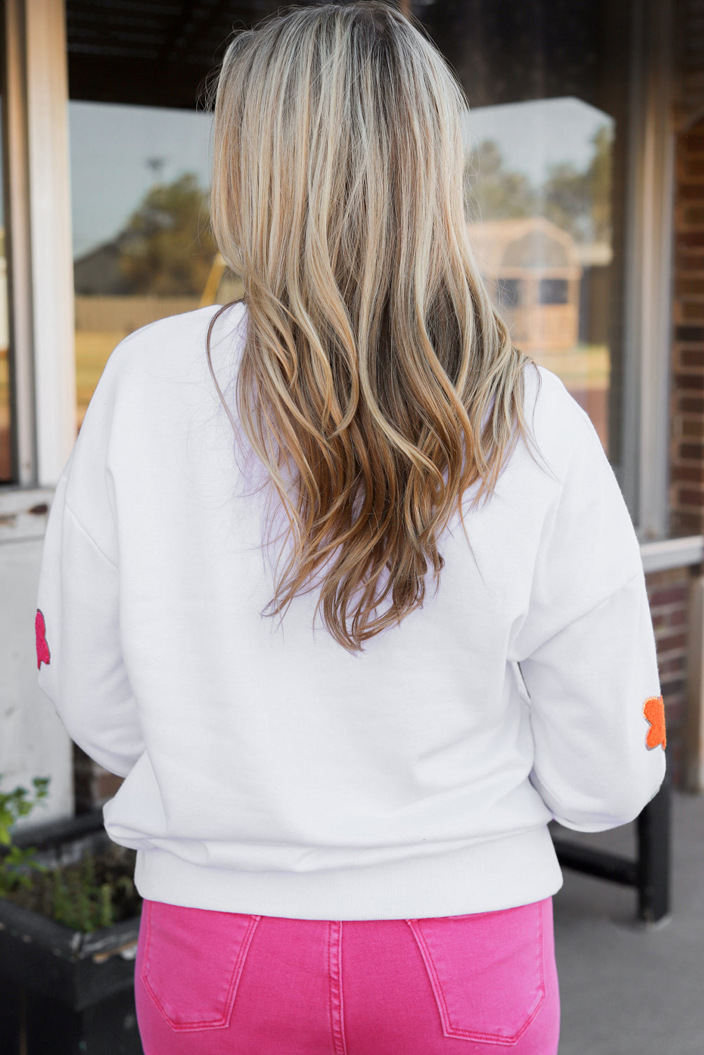 A person poses in the White Howdy Glitter Chenille Patch Graphic Casual Sweatshirt, adorned with colorful star patterns and the letters "WD." She wears light denim shorts and has shoulder-length blonde hair.