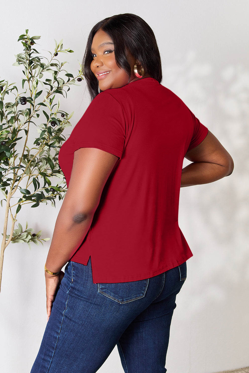 A person with wavy hair, wearing the Basic Bae Full Size Round Neck Short Sleeve T-Shirt in bright pink and beige pants, stands near a plant, smiling and looking to the side. The outfit embodies a basic style and offers slightly stretchy comfort, ideal for those who value effortless elegance.