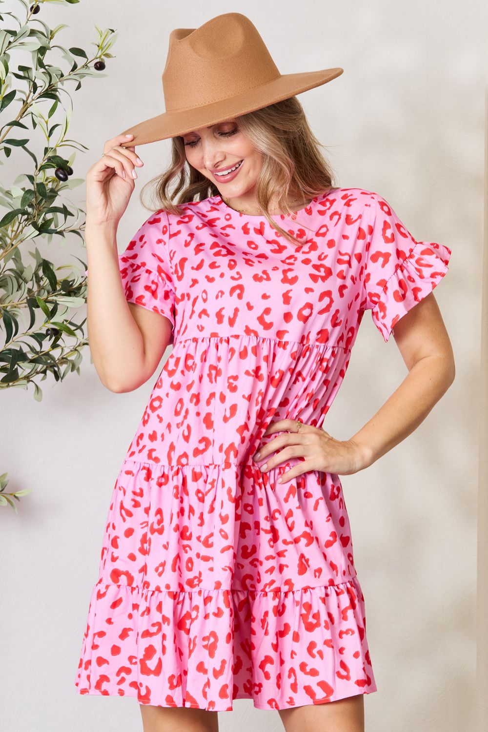 A woman wearing the Double Take Short Flounce Sleeve Tiered Dress in pink and red leopard print, complete with a ruffled hem and short flounce sleeves, smiles as she adjusts her brown hat. She stands near a plant, embodying the perfect boho-chic look.