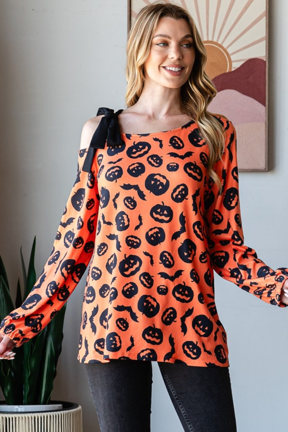 A woman smiles while modeling the Heimish Pumpkin Print One Shoulder T-Shirt, featuring an off-shoulder, long-sleeve design that is perfect for fall season fashion. She stands indoors with a plant and wall art in the background.