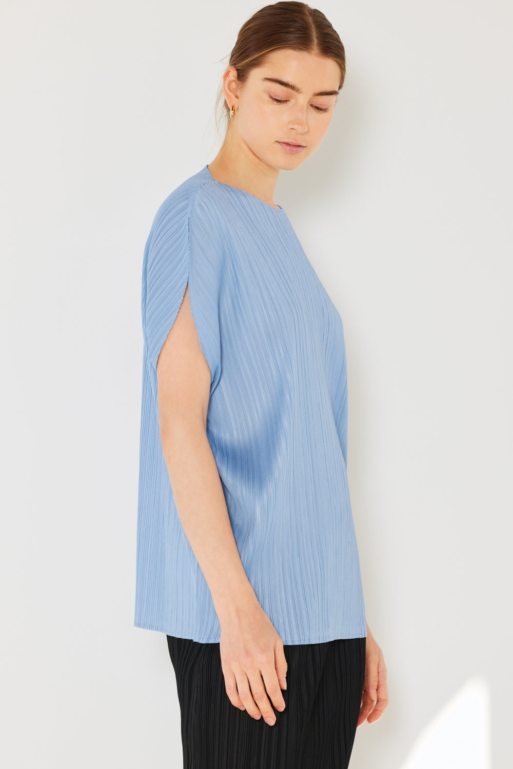 A person wearing the Marina West Swim Rib Pleated Oversized Dolman Sleeve Top in a fashionable light blue shade, paired with a brown belt and black skirt, stands against a plain background.