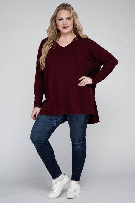 A woman with long, blonde hair wears a Plus Dolman Sleeve V-Neck Side Slit Hi-Low Hem Top and blue jeans, posing with one hand on her hip and smiling at the camera.