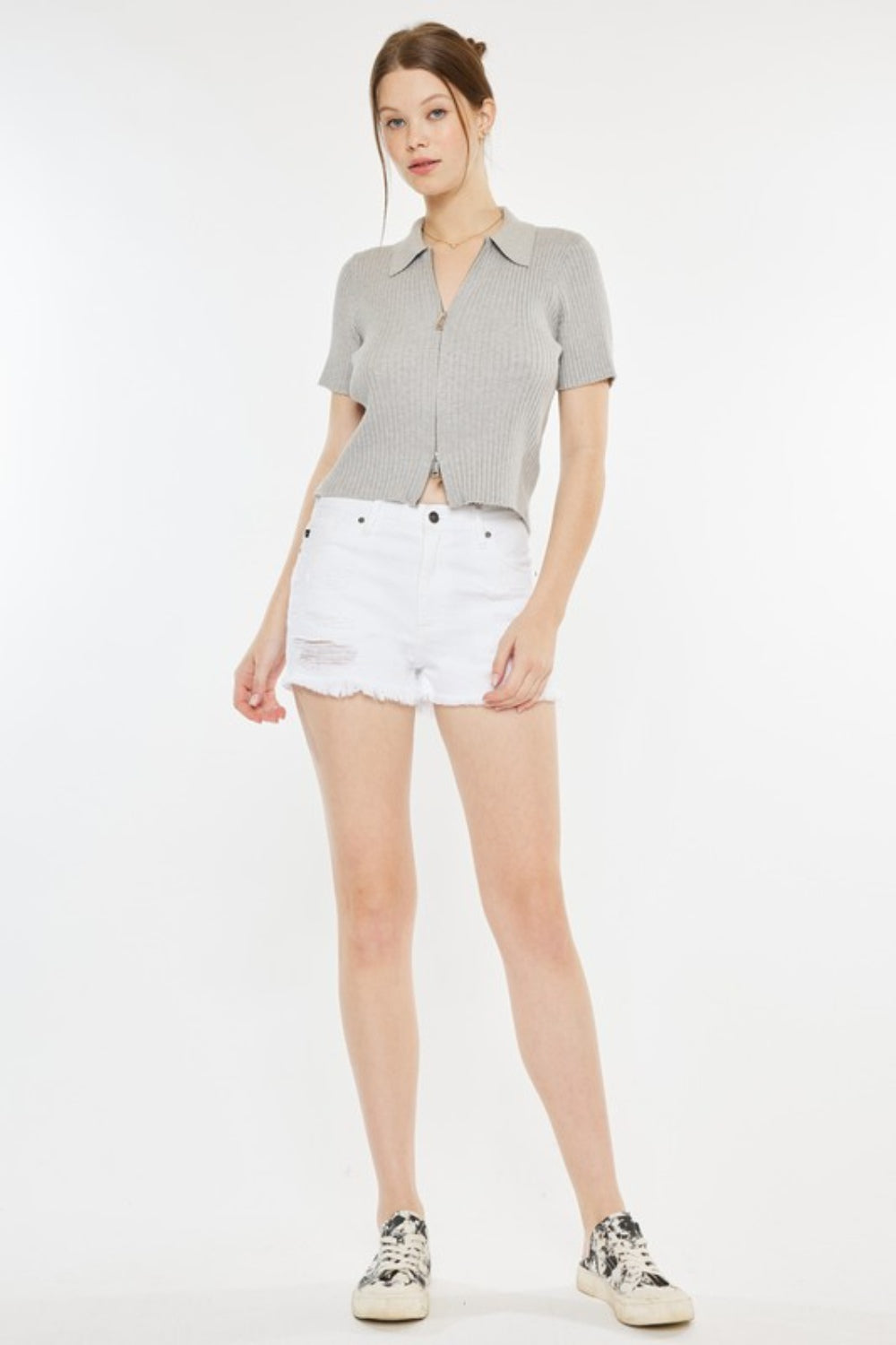 A person stands against a plain background, wearing a light grey short-sleeve top, Kancan Raw Hem Distressed Denim Shorts in white, and white slip-on shoes with black patterns.