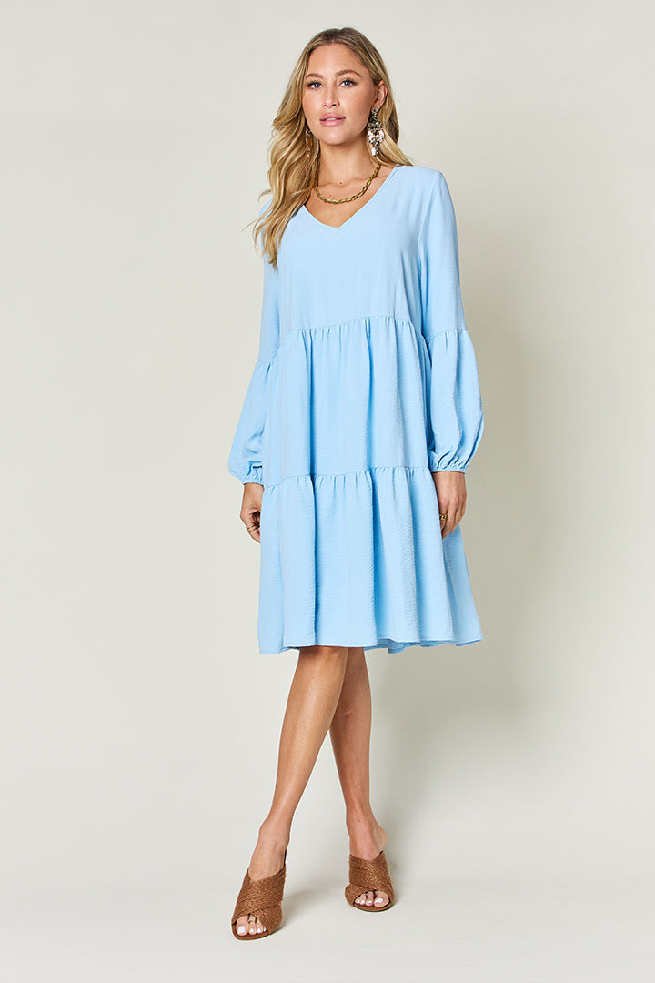 A woman stands wearing the Double Take Full Size V-Neck Balloon Sleeve Tiered Dress with Pockets in pink. Crafted from high stretch yarn, this dress combines comfort and style effortlessly. She has long, wavy hair and accessorizes with hoop earrings and a necklace. This imported garment is easy to care for – simply machine wash cold to keep it looking fresh.