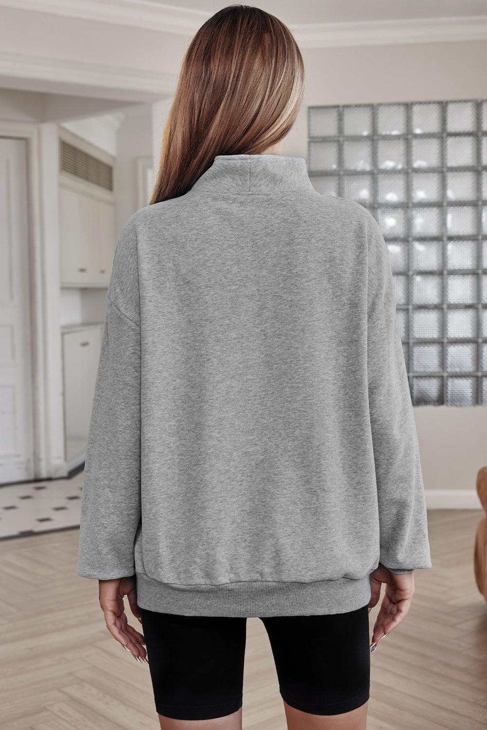 A person with long hair is standing indoors, facing away, wearing a Gray Zipped Funnel Neck Kangaroo Pocket Sweatshirt and black shorts. The background includes a kitchen area and a glass block wall.