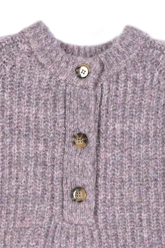 A person is wearing the Melange multicolor sweater top, characterized by textured pink fabric and featuring raglan sleeves along with large buttons, paired with light blue jeans, standing against a plain background.