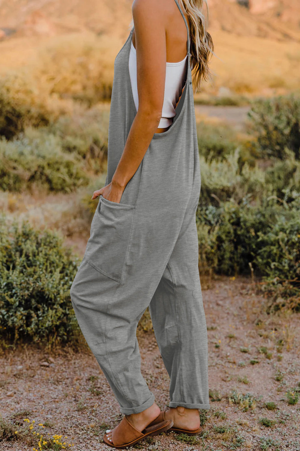 Person wearing a Double Take Full Size V-Neck Sleeveless Jumpsuit with Pockets and sandals, walking outdoors on a dirt trail with bushes in the background.