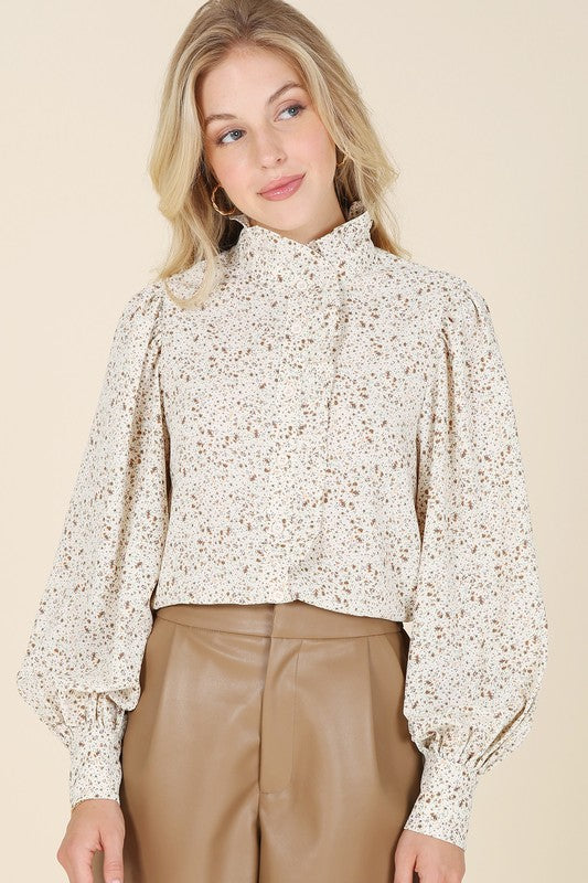 A woman wearing a stand collar floral frill blouse with puff sleeves and tan pants is standing against a beige background, looking to the side.