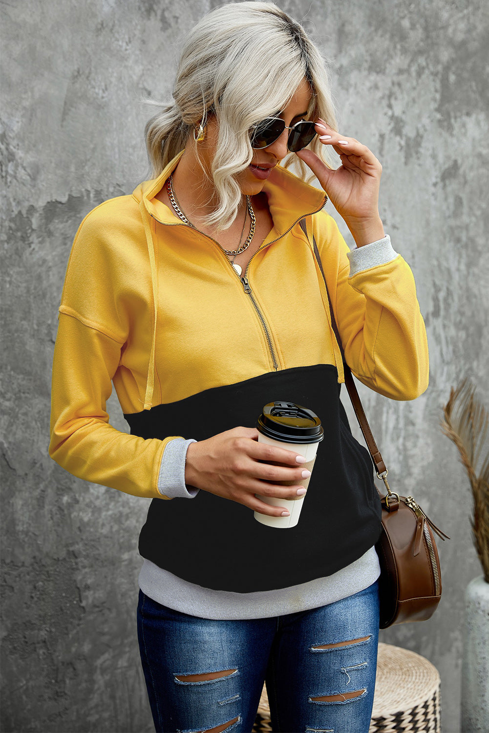 A person with long, light hair in a ponytail, wearing a Black Zipped Colorblock Sweatshirt with Pockets, blue jeans, and glasses, is holding a white cup and a brown handbag, shown from behind against a gray wall.