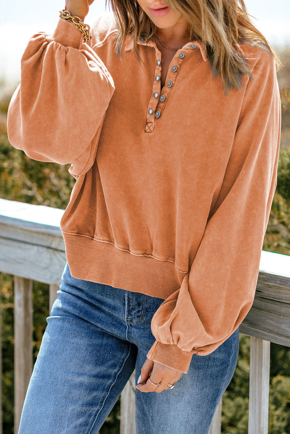 A person wearing an orange washed snap buttons lantern sleeve pullover sweatshirt and blue jeans stands with one hand on their hip, facing away from the camera, leaning slightly on a wooden rail outdoors.