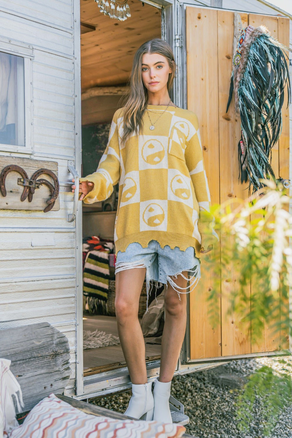 A woman wearing the And The Why Checkerboard Sweater with a Yin Yang pattern and distressed denim shorts stands in a doorway.