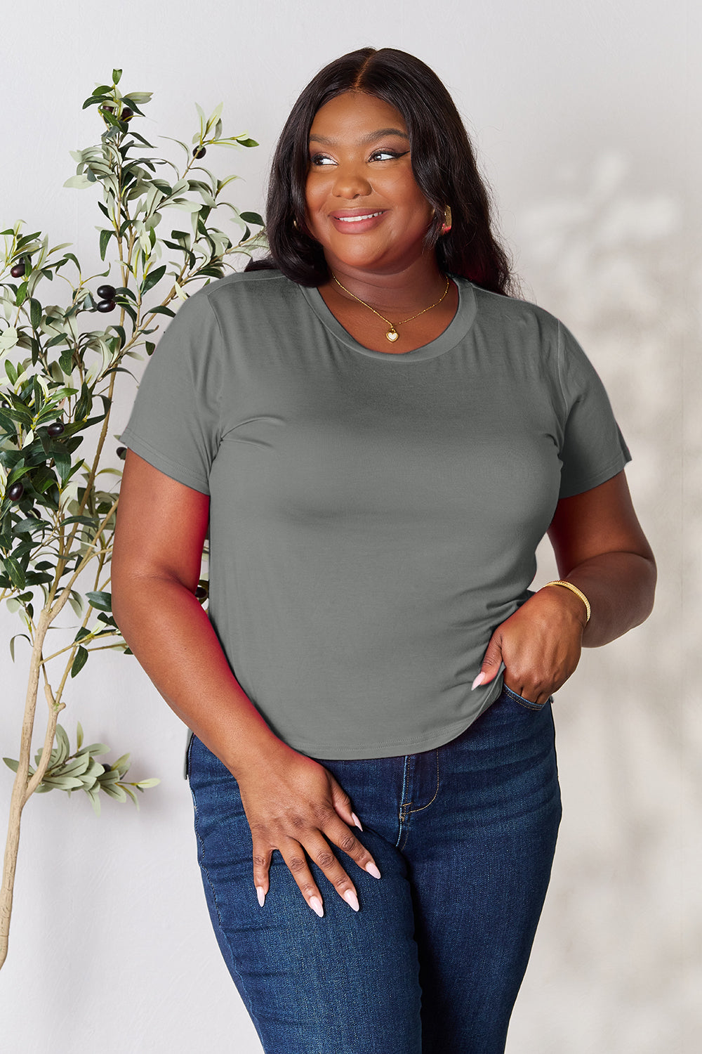 A person with wavy hair, wearing the Basic Bae Full Size Round Neck Short Sleeve T-Shirt in bright pink and beige pants, stands near a plant, smiling and looking to the side. The outfit embodies a basic style and offers slightly stretchy comfort, ideal for those who value effortless elegance.