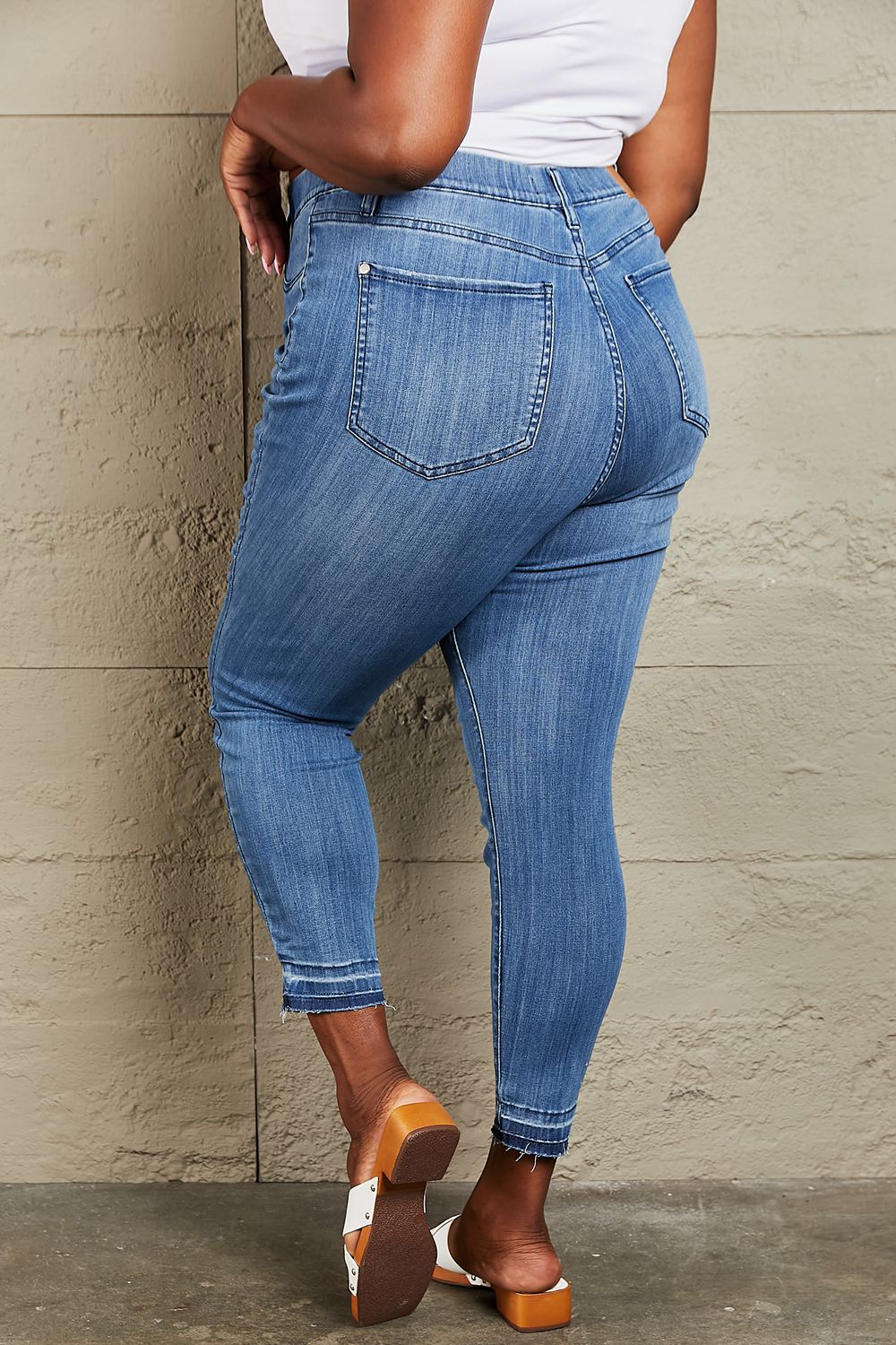 Person wearing Judy Blue Janavie Full Size High Waisted Pull On Skinny Jeans with release hem detailing and white sandals stands against a textured wall.