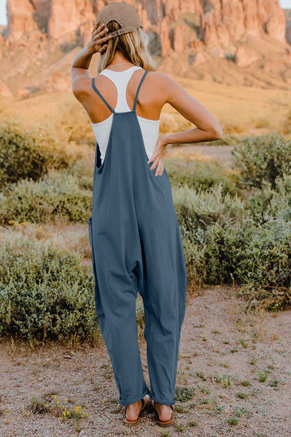 Person wearing a Double Take Full Size V-Neck Sleeveless Jumpsuit with Pockets and sandals, walking outdoors on a dirt trail with bushes in the background.