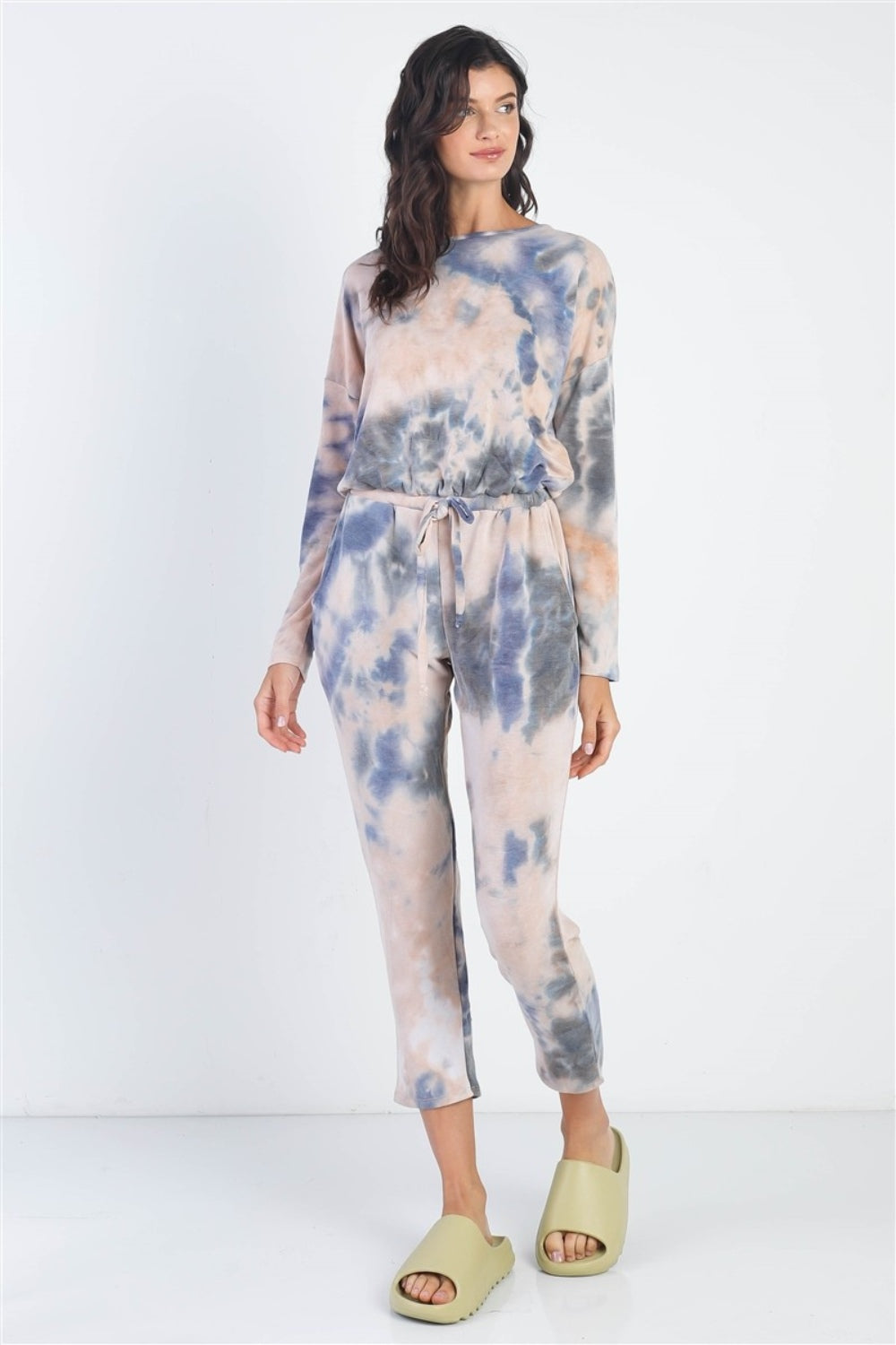 Dressed in a Cherish Apparel Tie-Dye Round Neck Long Sleeve Jumpsuit and green sandals, a person strikes a street-style pose against a plain white background.