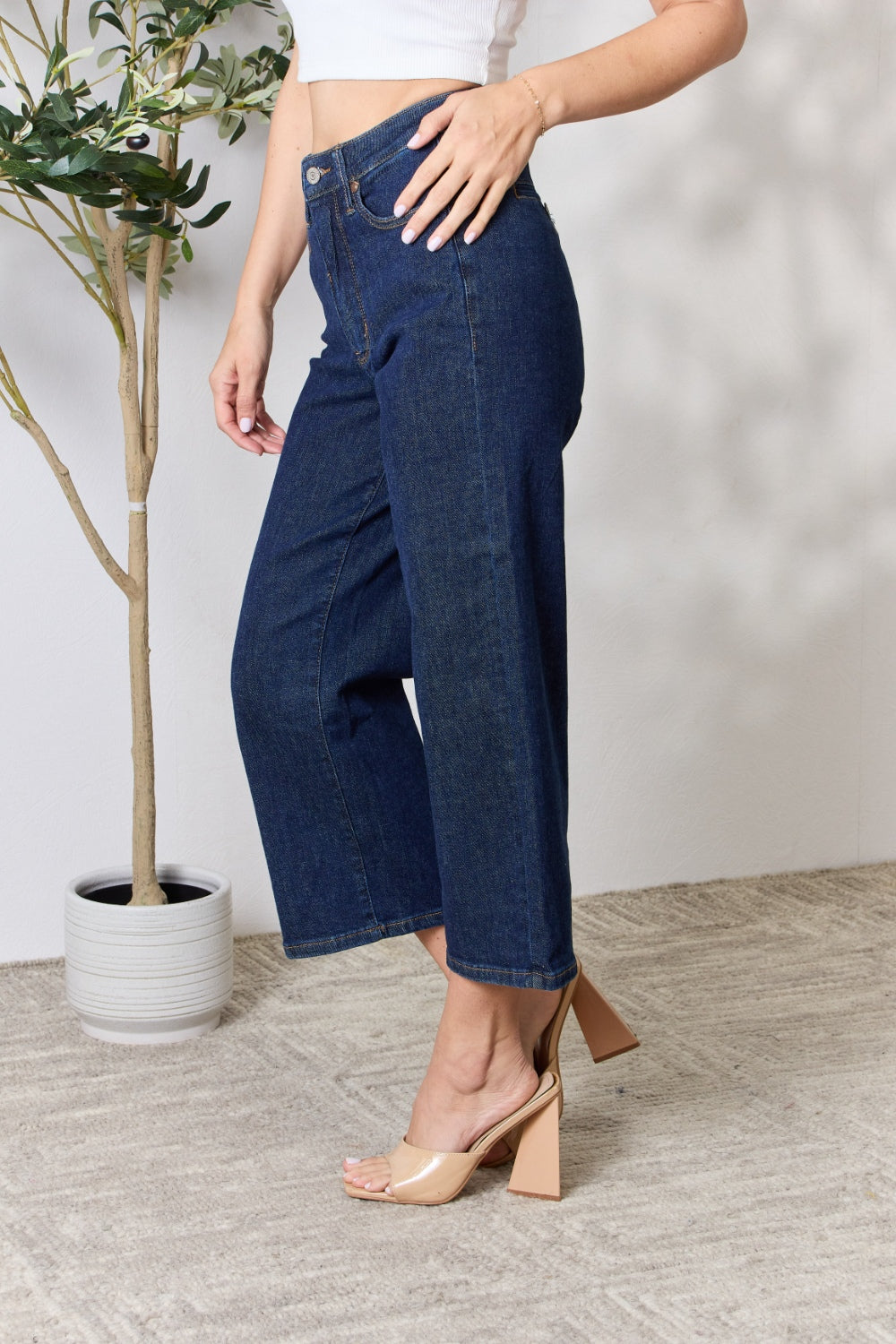 A person wearing the Judy Blue Full Size High Waist Cropped Wide Leg Jeans, which are renowned for their tummy control design, and nude block heel sandals stands near a potted plant.