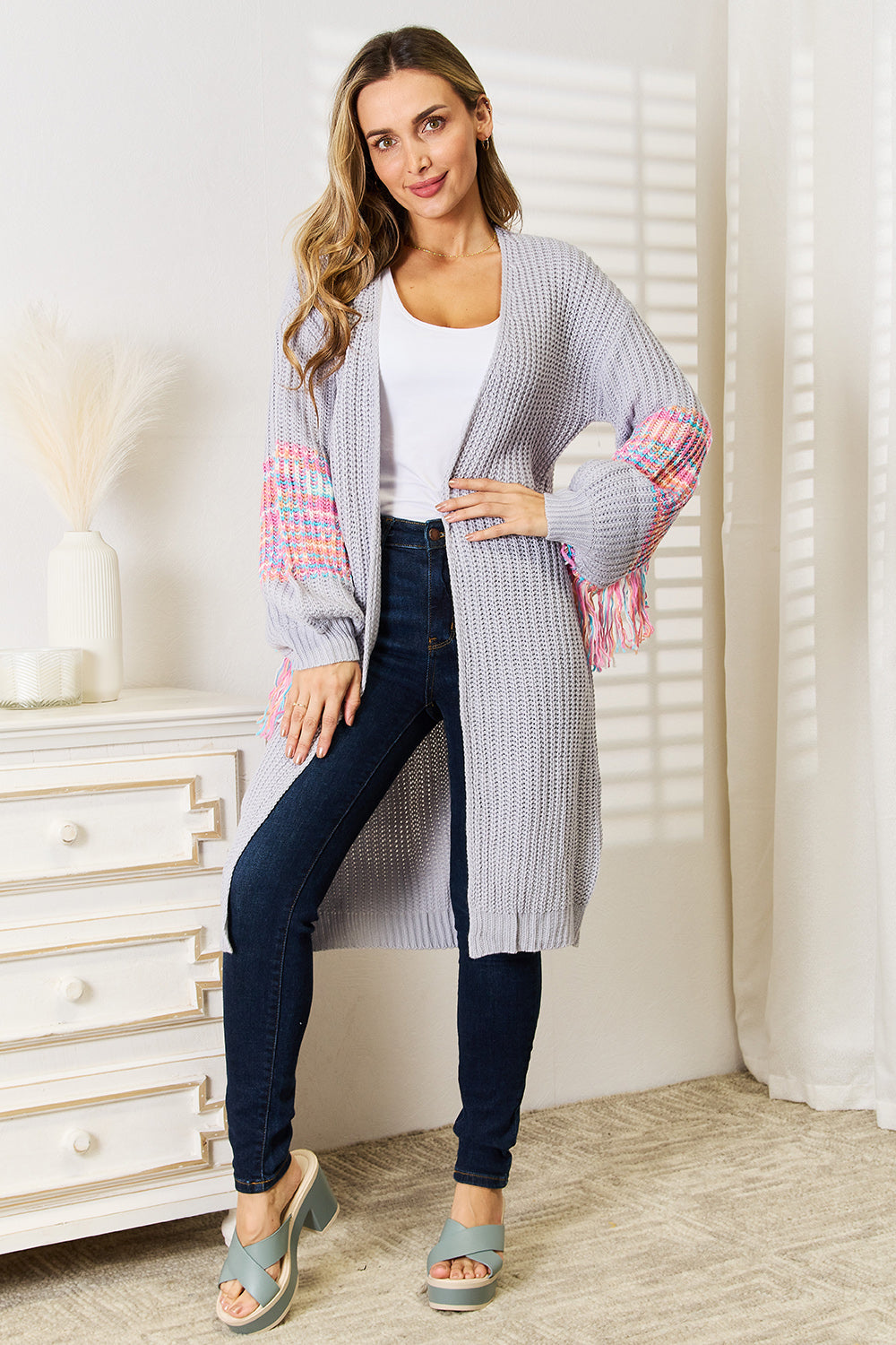 A woman poses indoors wearing the Woven Right Fringe Sleeve Dropped Shoulder Cardigan, a cozy, light gray piece featuring colorful tasselled sleeves. She pairs it with a white top and dark jeans.