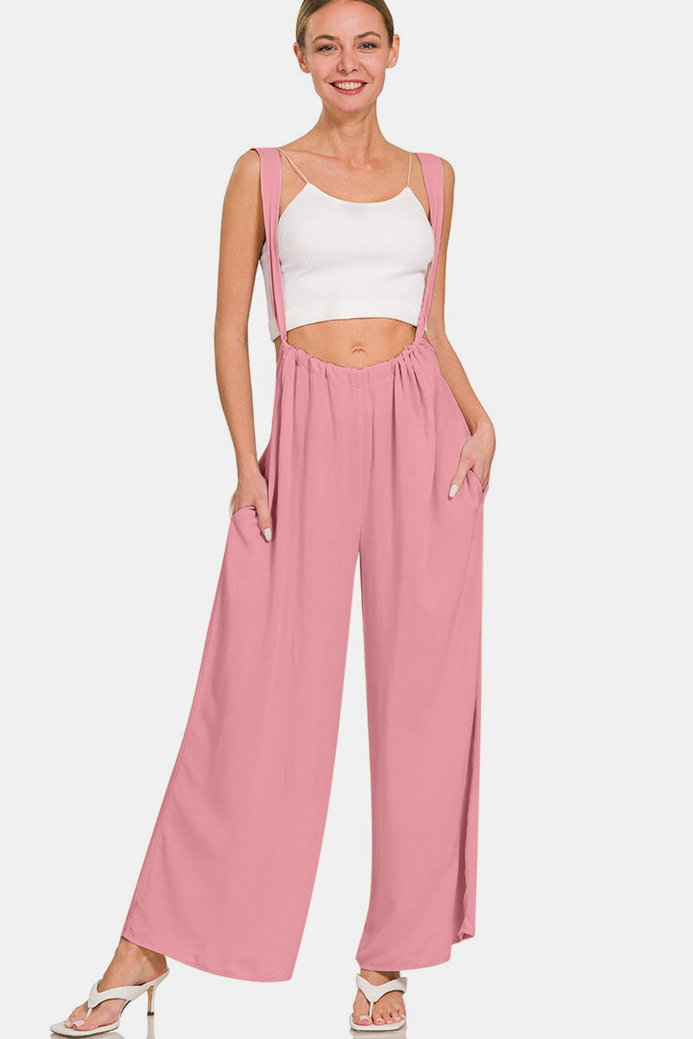 A woman is smiling in her fashion-forward style, donning the Zenana Pocketed Wide Strap Wide Leg Overalls in pink along with a white crop top. She has her hands in the overall's pockets and complements the look with white high-heeled shoes.