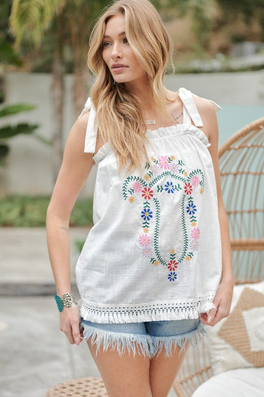 A person wearing an Embroidered Sleeveless Ruffle Tank Top and denim shorts stands outdoors near a wicker chair, adjusting their hair.